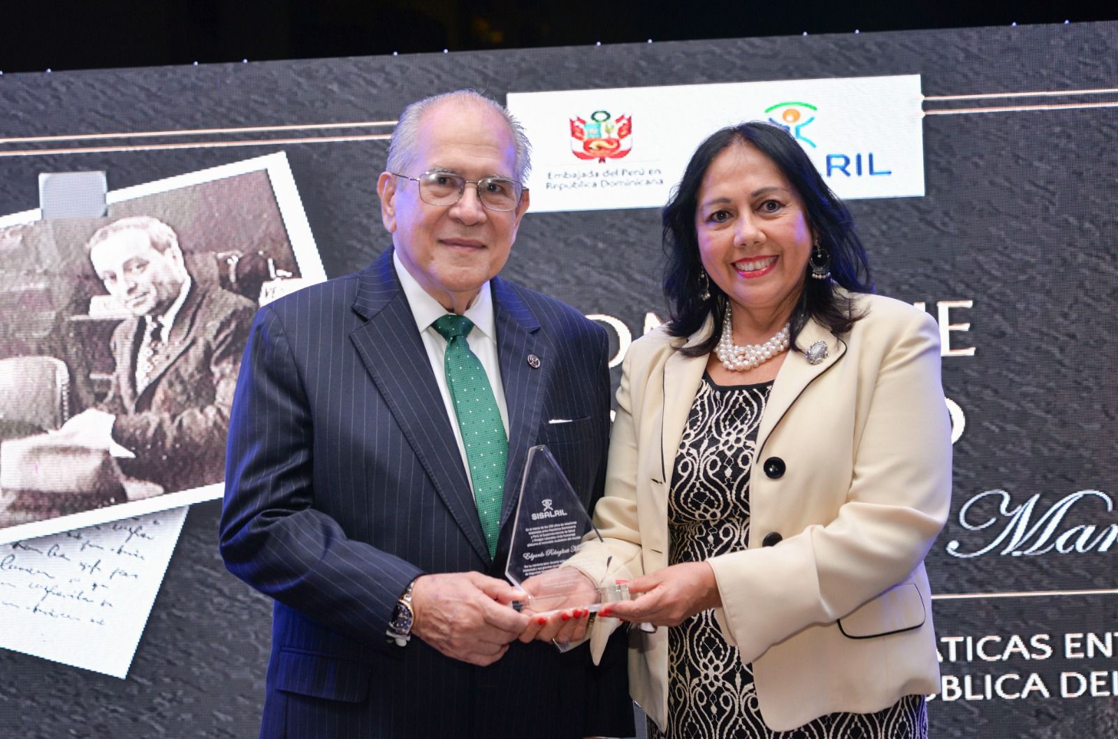 El Dr. Jesús Feris Iglesias, superintendente de la SISALRIL, entrega el reconocimiento a la Sra. Embajadora del Perú en el país, María Cecilia Rozas Ponce de León, quien a nombre de la familia Rebagliati Martins recibió el reconocimiento otorgado por la SISALRIL. (LaSeguridadSocial.Do)